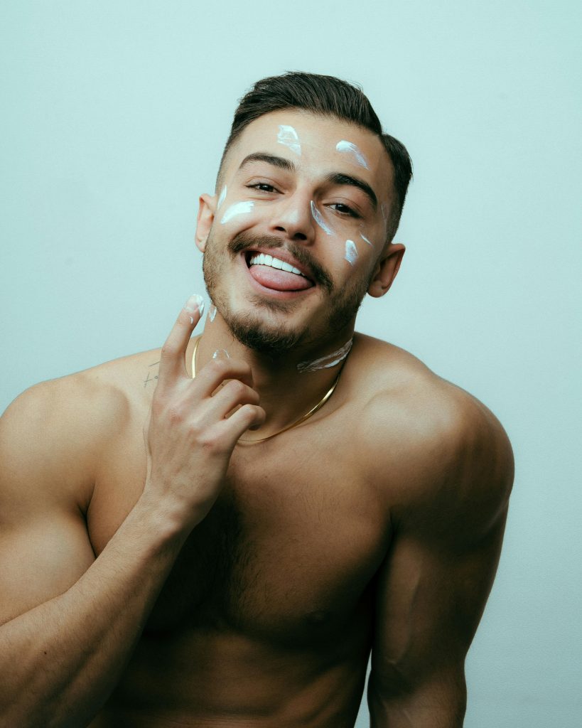 Man smiling while applying facial skincare lotion against light background.